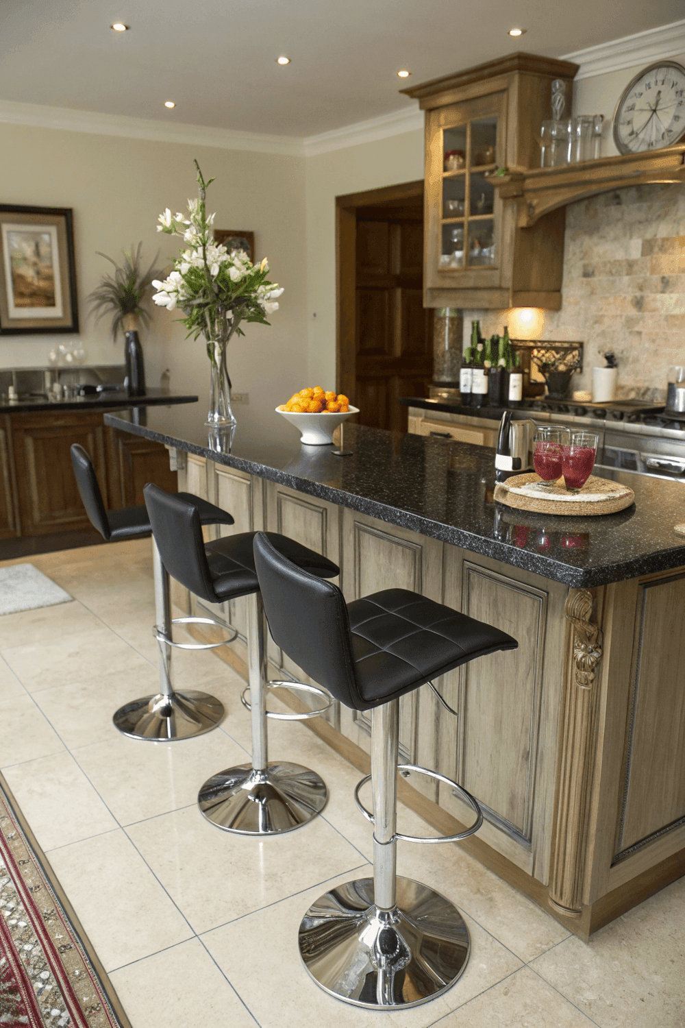 13b840804b3d4aec90a76e915b87fa72 A photo of a modern kitchen with a sleek marble topped counter and black leather bar stools The kitchen has wooden cabinetry a stone accent wall and a variety of accessories including a vase with fresh white flowers a bowl of citrus fruits wine bottles and a warm inviting atmosphere The lighting is soft and ambient The photo is taken from a perspective that provides a view of the bar stools counter and some background elements The image is a high resolution photograph