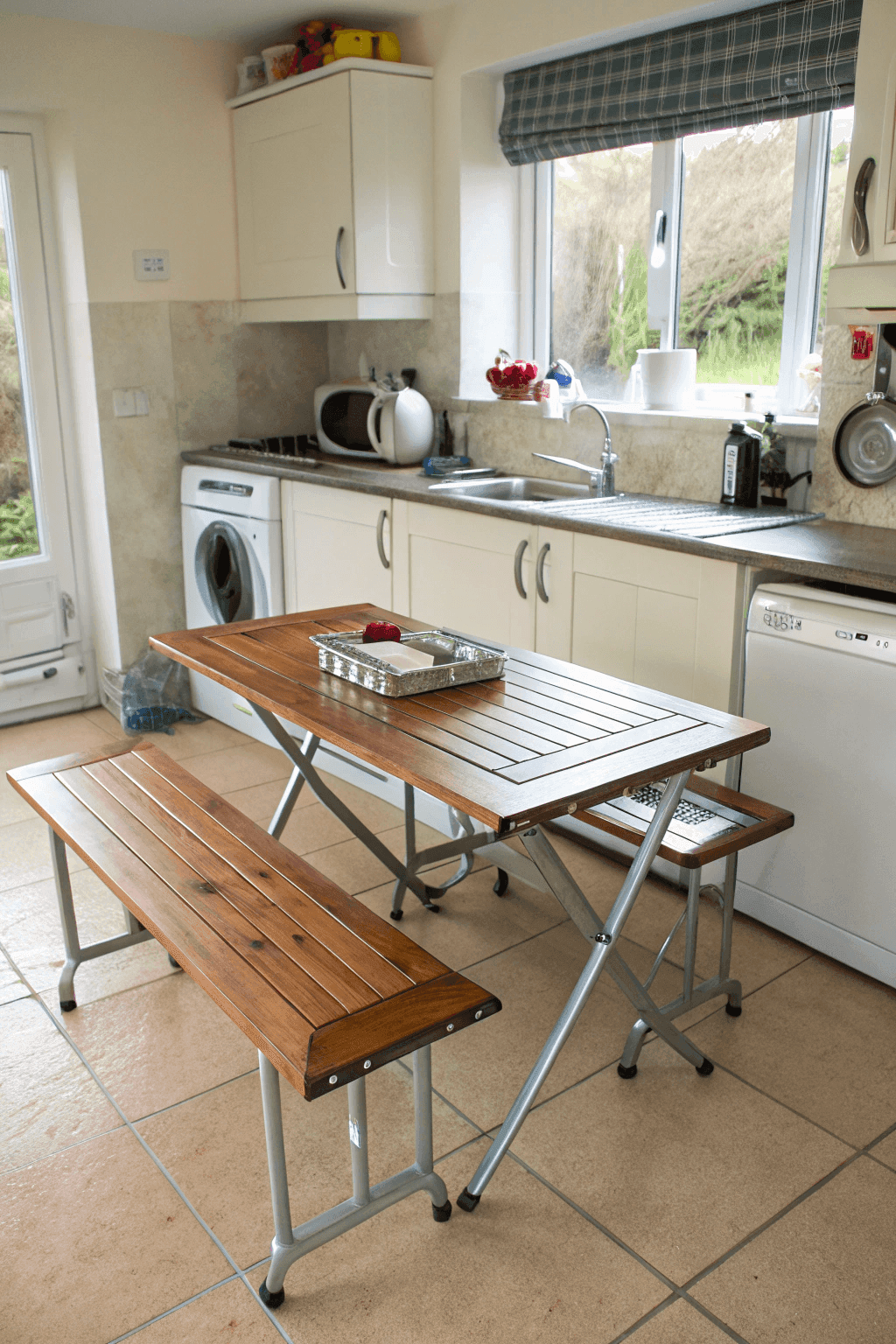 89a4e130b40e42b1b8aafd788023d0ec A photo of a kitchen with a wooden folding table and bench seating in the foreground a kitchen sink a kettle a kitchen countertop a window and a washing machine The kitchen has a modern functional design with white cabinets and a view of greenery outside The wooden table and bench provide a warm natural touch The image is a photograph with natural lighting coming through the window creating a soft and welcoming ambiance The colors are natural and muted