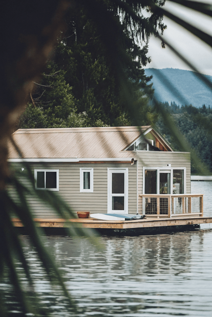 The dock house A unique tiny house floating on a dock surrounded by clear blue water Paddleboards are tied to the side and the wraparound deck provides panoramic views of the lake and distant mountains