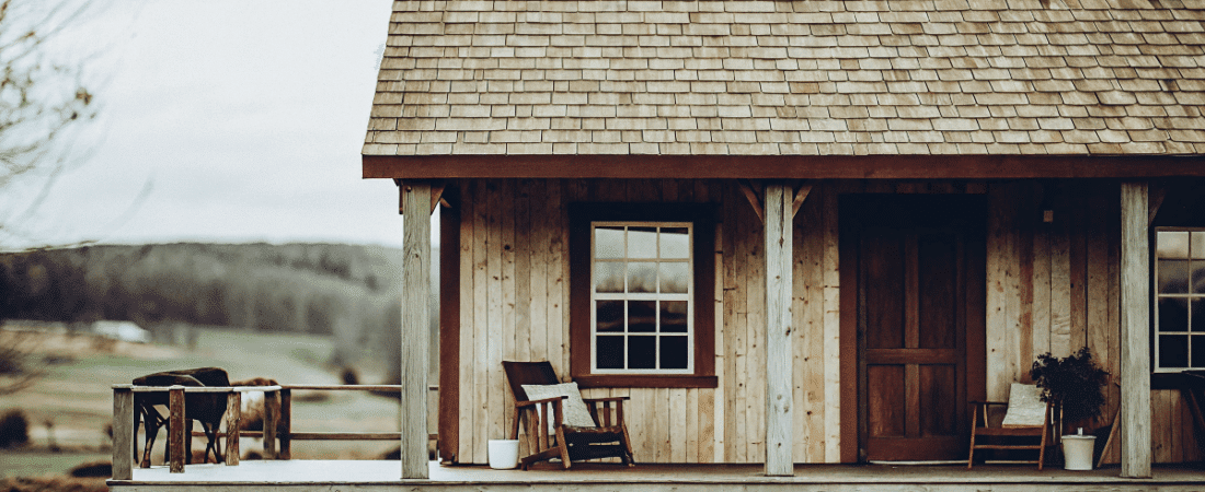 A rustic wooden cabin with a porch extending from the structure featuring rocking chairs potted plants and a simple traditional design The cabin is constructed with a combination of wooden beams and a sloped roof made of shingles The scene is set in a rural area with a vast open field and gentle hills visible in the background The overcast sky adds a soft diffused light to the overall scene The image style is a photo with a muted color palette