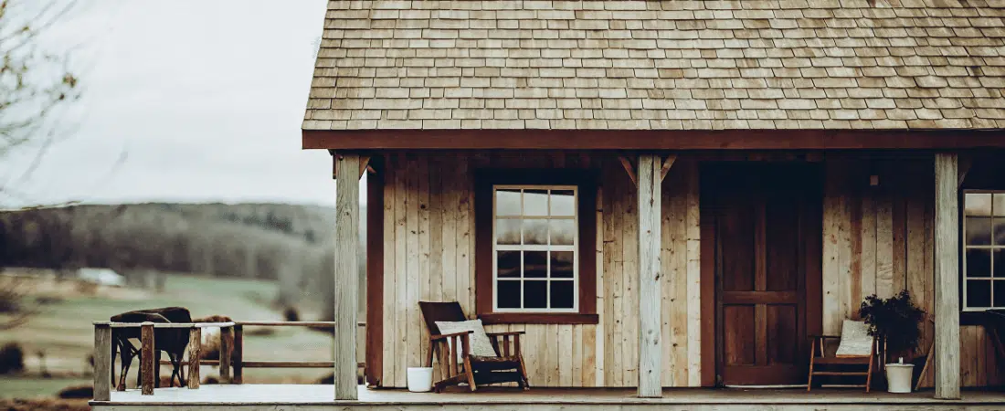 A rustic wooden cabin with a porch extending from the structure featuring rocking chairs potted plants and a simple traditional design The cabin is constructed with a combination of wooden beams and a sloped roof made of shingles The scene is set in a rural area with a vast open field and gentle hills visible in the background The overcast sky adds a soft diffused light to the overall scene The image style is a photo with a muted color palette