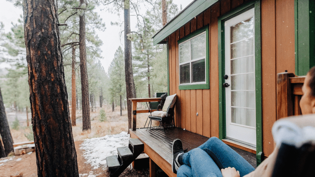 Lake tahoe A cozy tiny house tucked among towering pine trees, featuring a private deck overlooking a shimmering lake. The scene is tranquil, with a light dusting of snow on the ground, evoking a serene winter getaway.