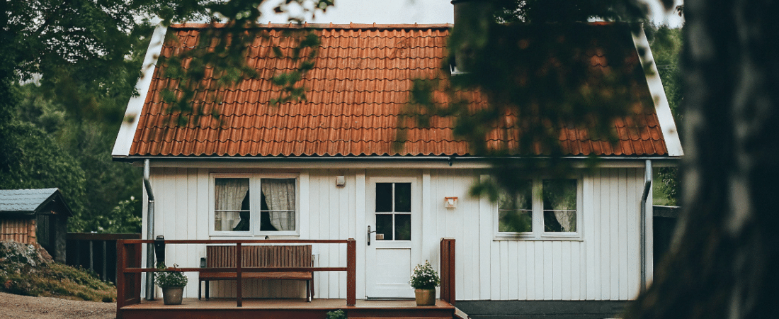 A photo of a quaint cottage nestled in a wooded area The cottage predominantly white with a distinctive orange tiled roof features a front porch with a wooden railing and several potted plants The architecture is simple and rustic with a traditional design The photo is taken from a vantage point slightly obscured by the foliage of nearby trees providing a sense of depth and a feeling of being enveloped by nature The image style is a photograph