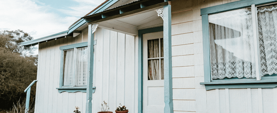 A photograph of a quaint cottage style house with a predominantly white exterior accented with teal colored trim