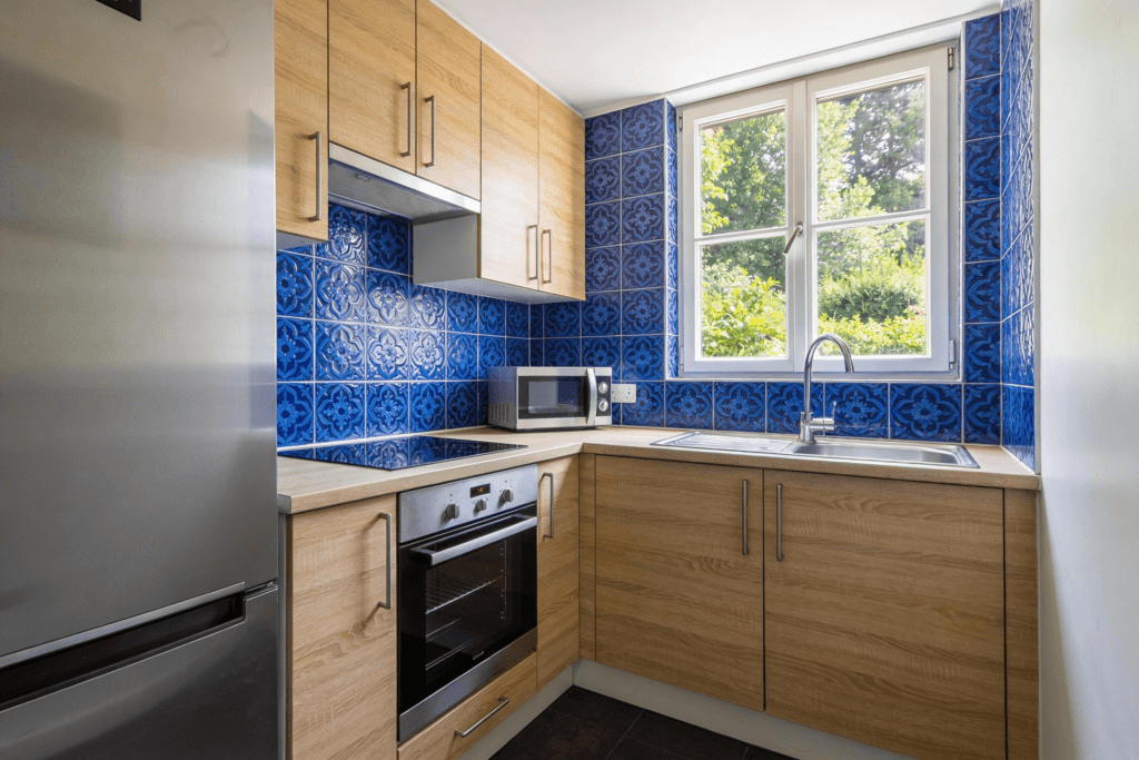 The faux tile backsplash Photograph of a compact modern kitchen featuring vibrant blue patterned tiles light wood cabinetry stainless steel appliances and a window overlooking greenery