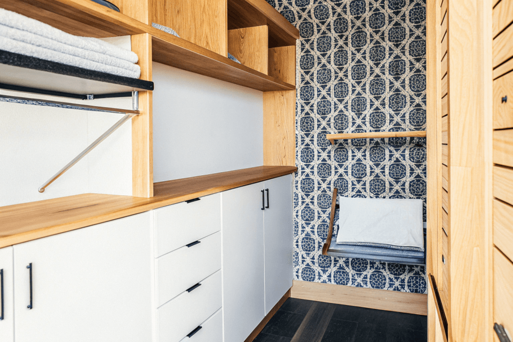 Cozy upscale laundry room with white oak cabinetry wood floors ceiling mounted drying rack built in ironing board and patterned wallpaper