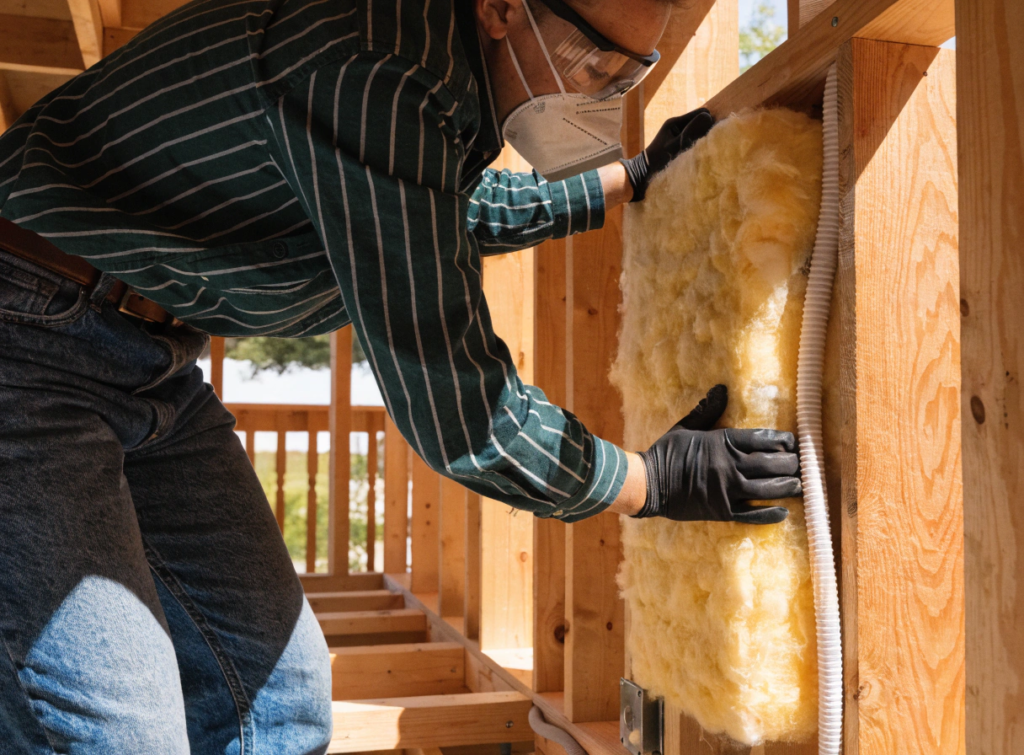 Person with mask Person wearing safety goggles and a mask installs yellow fiberglass insulation between wooden studs in a building frame