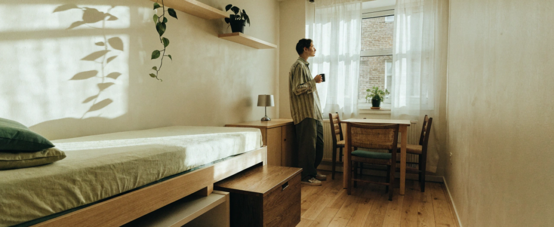 Small studio apartment A bright welcoming studio apartment shot from a corner to show the whole room at once Roughly 400 square feet with a fold down bed a small dining nook by a large window and a couple of leafy plants on floating shelves Warm morning light pours in through sheer curtains A person in casual clothes stands near the window holding a coffee mug looking relaxed and at home The color palette is soft light wood cream walls muted green accents The mood should feel calm roomy and lived in rather than staged Wide angle framing natural light no clutter